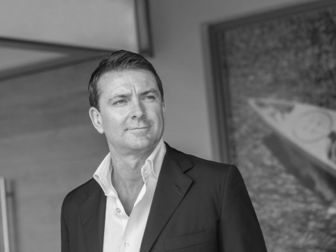 A man in a suit jacket and open-collared shirt stands indoors, looking slightly to the side with a neutral expression. The image is in black and white, with a blurred background featuring artwork.