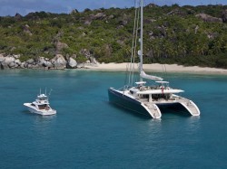 A large catamaran and a smaller motorboat are anchored in clear blue water near a tropical beach with dense green vegetation and rocky hills in the background.