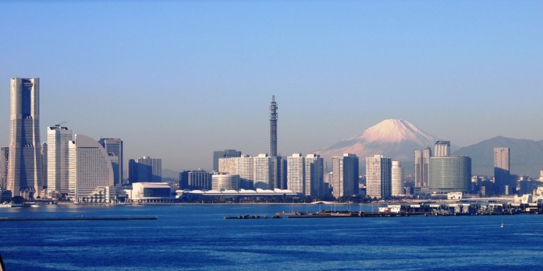 A city skyline with modern high-rise buildings by the waterfront, with snow-capped Mount Fuji visible in the background under a clear blue sky.