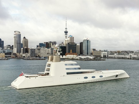 A modern white luxury yacht sails in a harbour with a city skyline and tall buildings, including a prominent tower, under a cloudy sky in the background.