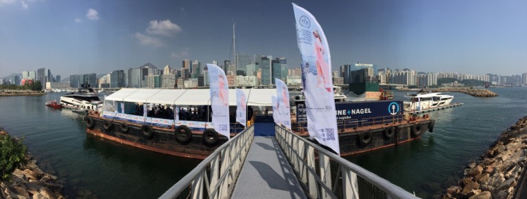 A moored barge with a white marquee and banners sits on the water near a city skyline with many high-rise buildings under a clear blue sky. A walkway leads from the foreground to the barge.