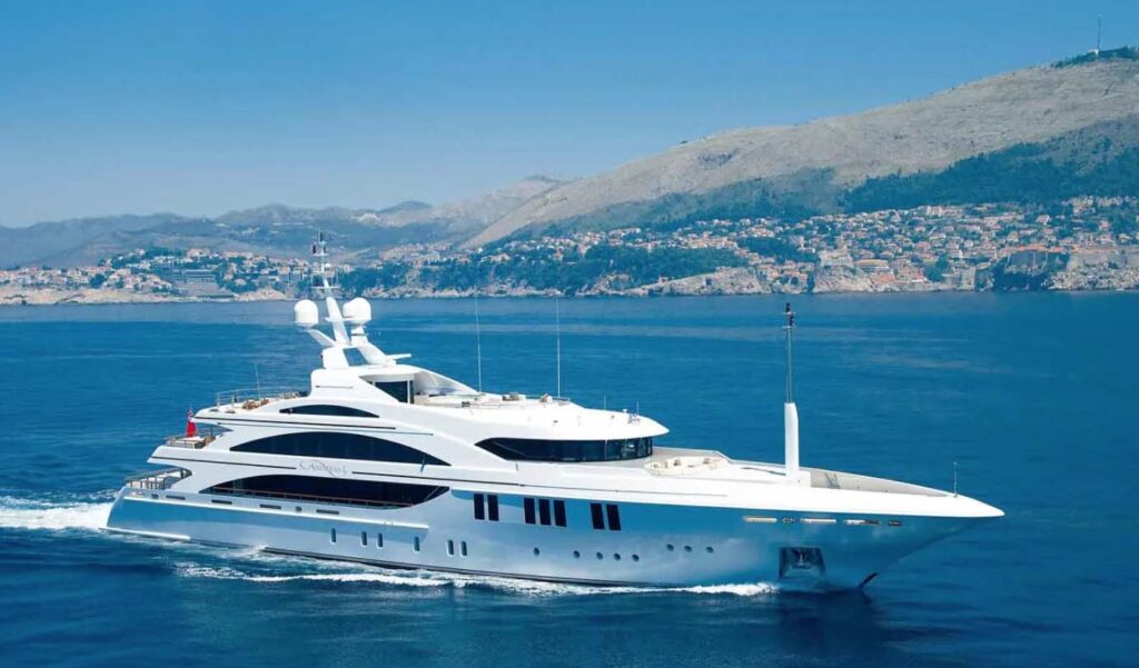 A large white luxury yacht sails on calm blue water with hilly coastline and a town visible in the background under a clear sky.