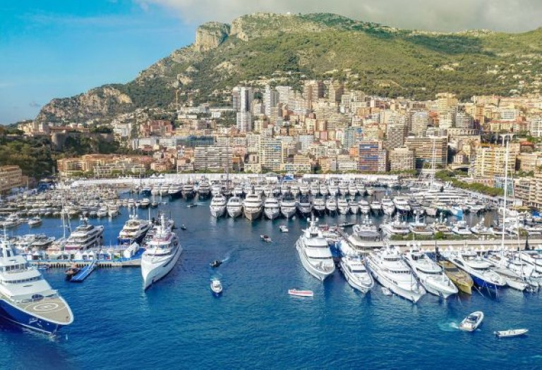 Aerial view of a large marina filled with numerous yachts and boats, with a city of tall buildings and green hills rising in the background under a partly cloudy sky.