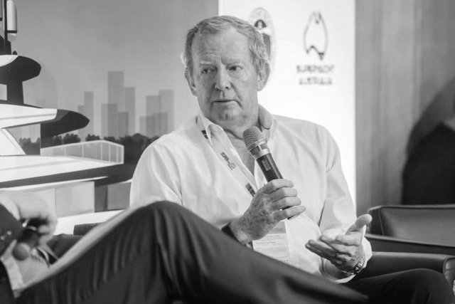 A man in a white shirt is speaking into a microphone while seated. He gestures with his left hand during what appears to be a panel discussion or interview. The setting is indoors, with a backdrop and logos behind him.
