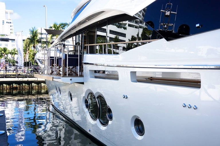 A modern white yacht moored at a marina, with its shiny exterior reflecting in the water. Palm trees and tall buildings are visible in the background under a clear sky.