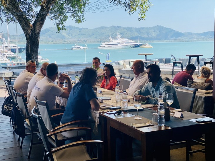 A group of people converse around a table at a waterfront restaurant, with yachts and mountains visible across a sunny blue harbour in the background.