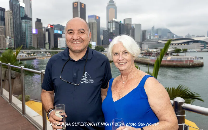 A smiling man and woman pose together on a waterfront terrace with a city skyline and harbour in the background. The man wears a navy shirt and the woman a blue dress. Text at the bottom reads, APSA ASIA SUPERYACHT NIGHT 2024. photo Guy Nowell.