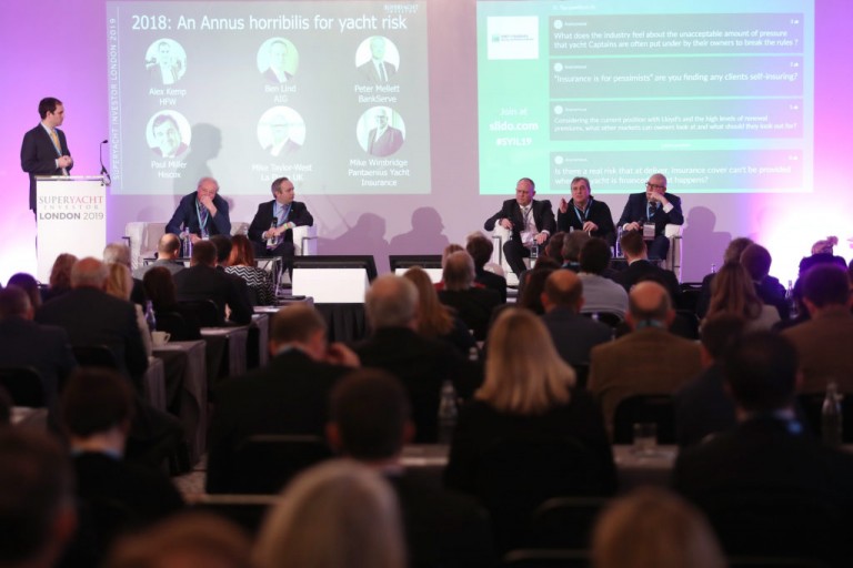 A man stands at a lectern while five panellists sit on stage discussing yacht risk at the Superyacht Conference London 2018. The audience is seated, and slides with discussion topics are displayed in the background.