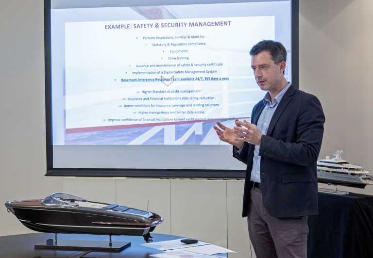 A man stands next to a model yacht, giving a presentation on yacht safety and security management. Behind him is a large screen displaying detailed information and example points about the topic.