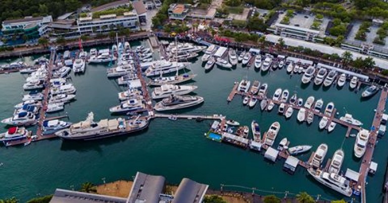 Aerial view of a marina filled with numerous yachts and boats moored at piers, surrounded by waterfront buildings and greenery. The water is calm and the scene is bustling with activity.