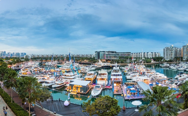 A vibrant marina filled with numerous yachts and boats moored at piers, surrounded by palm trees, waterfront buildings, and a lively crowd under a blue sky during dusk.