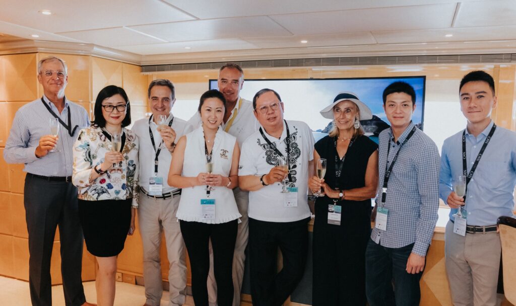 A group of nine people dressed in business or smart casual attire stand indoors, smiling at the camera and holding glasses, possibly toasting at a corporate or social event. Most have name badges around their necks.
