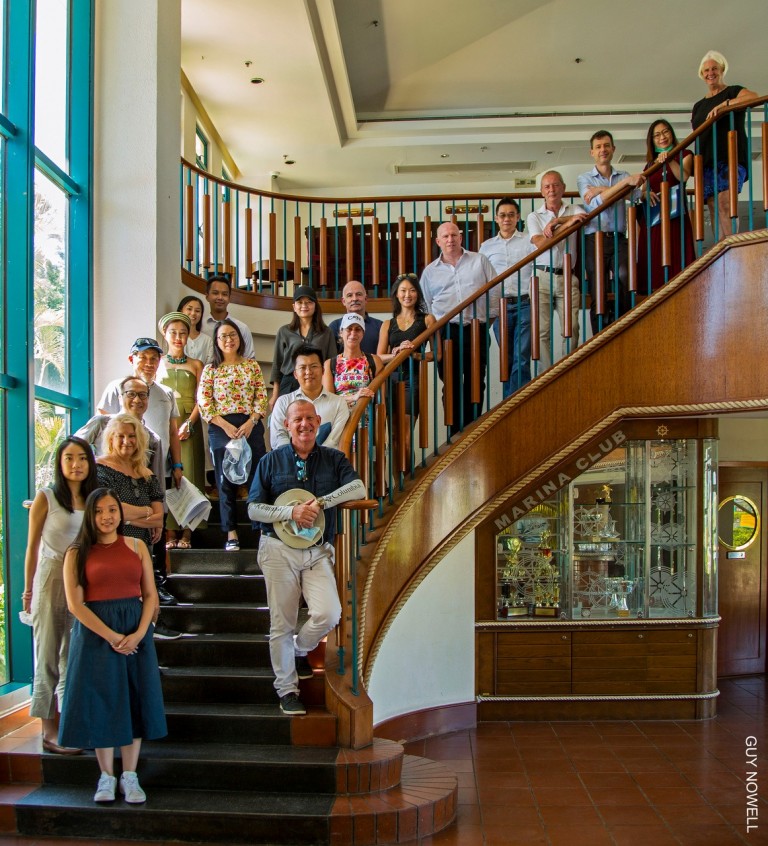 A group of people pose and smile on a curved staircase inside a building with large windows and a display cabinet; the group includes a mix of ages and is casually dressed.