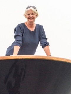 A smiling older woman with short grey hair wearing a blue top stands behind a curved wooden structure, with a light background.