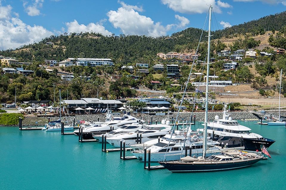 A marina with several luxury yachts and boats moored in turquoise water, set against a backdrop of green hills and buildings under a partly cloudy sky.