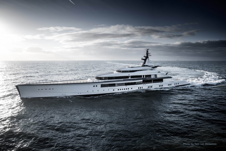 A sleek, modern white yacht sails across a calm, open sea under a partly cloudy sky, leaving a gentle wake behind it. The sun is low on the horizon, creating a dramatic light and shadow effect on the water.