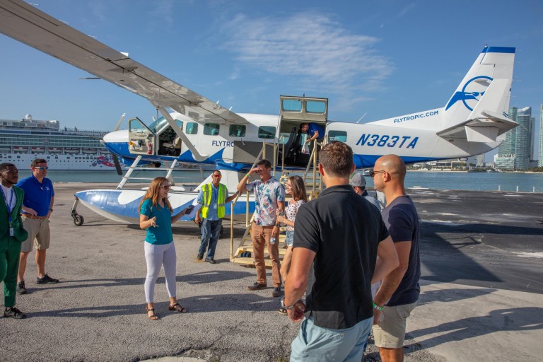 A group of people stands on a runway near a white and blue seaplane, talking and preparing to board. A cruise ship and city buildings are visible in the background under a clear sky.