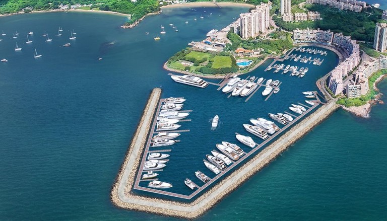 Aerial view of a marina with many yachts and boats moored, surrounded by stone breakwaters, with nearby residential buildings, green hills, and sailing boats on the surrounding blue water.