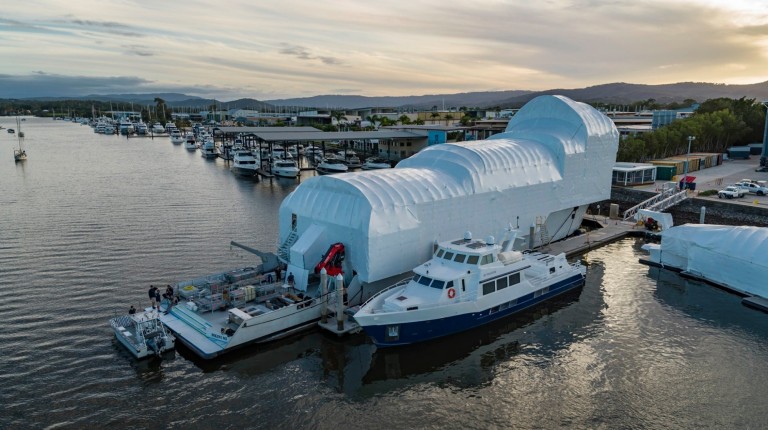 Aerial view of two boats moored beside a large, white, tent-covered structure at a marina, with several other boats and buildings visible along the waterfront under a cloudy sky.