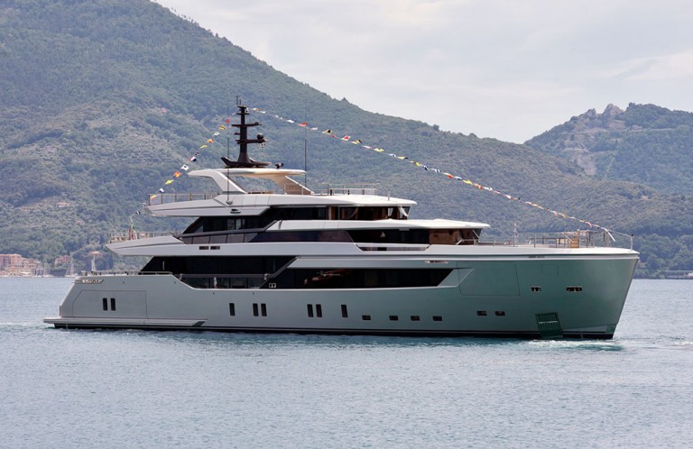 A modern luxury yacht with multiple decks floats on calm water, decorated with colourful flags. Green hills and cloudy sky form the backdrop.