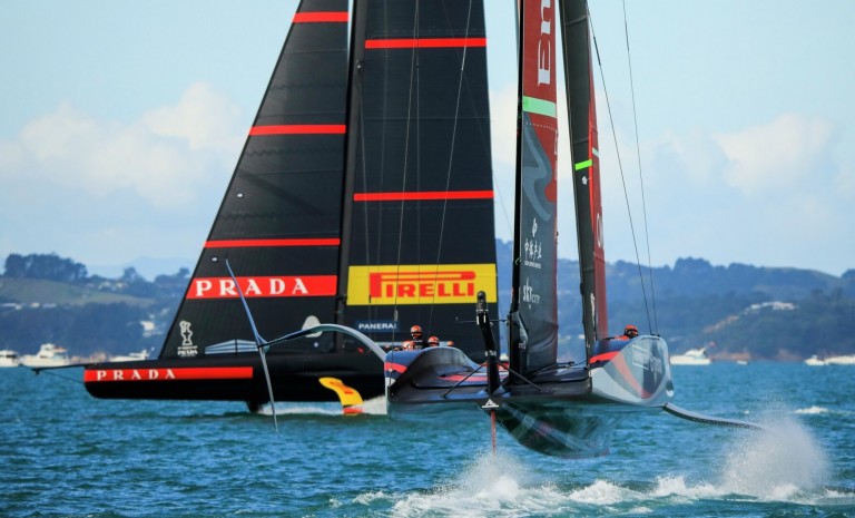 Two sleek racing yachts, featuring black sails with red PRADA and yellow PIRELLI logos, compete on blue water with distant boats and a hilly shoreline in the background. One yacht lifts partially above the water.
