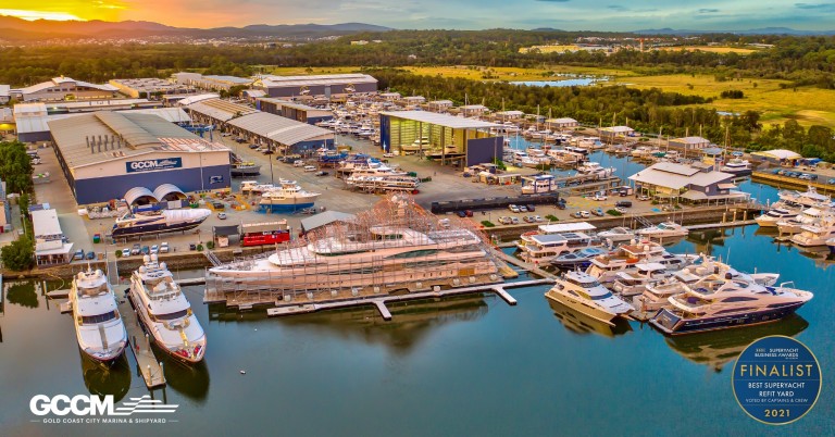 Aerial view of a large marina and shipyard with several yachts moored, buildings, and lush greenery in the background. The GCCM and award logos are visible in the bottom corners of the image.