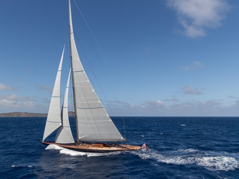 A sleek sailboat with tall white sails glides swiftly over deep blue ocean waves under a clear sky with scattered clouds. Land is visible in the distance on the left side of the image.