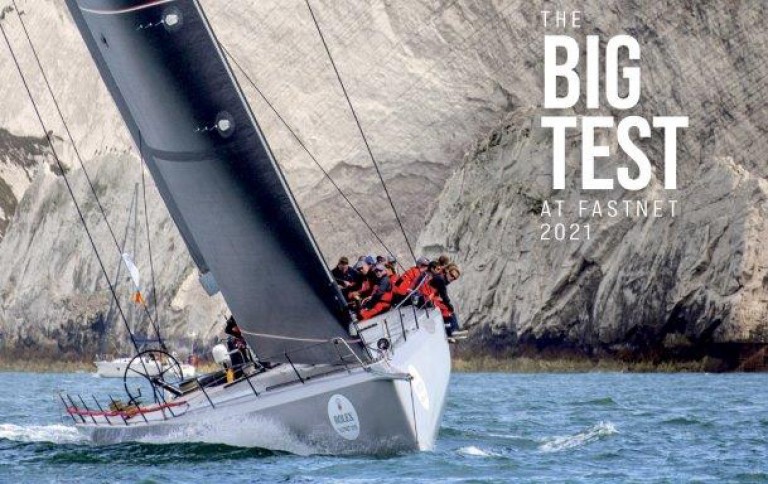 A sailboat with crew members in red jackets leans dramatically whilst racing near rocky cliffs. Large text reads The Big Test at Fastnet 2021. The water is choppy under a cloudy sky.