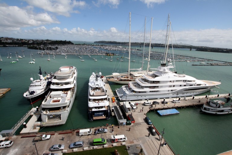 Aerial view of several large yachts moored at a marina on turquoise water, with more boats and a bridge in the background under a partly cloudy sky.