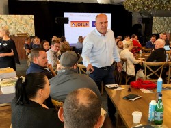 A man stands and speaks to seated attendees at a workshop or seminar. People are gathered around tables with notebooks and drinks. A presentation screen in the background displays the Jotun logo.