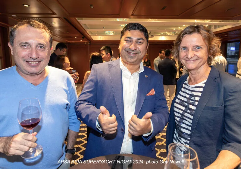 Three people smile at a social event; the man in the centre gives two thumbs up, while the others hold drinks. Other attendees mingle in the background inside a well-lit, elegant room.