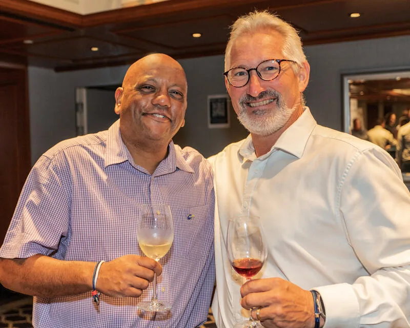 Two men smiling and posing together indoors, each holding a glass of wine. They are dressed in collared shirts and standing in a warmly lit room with wooden accents and a mirror in the background.