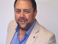 A man with short dark hair and a trimmed beard, wearing a light beige jacket over a blue shirt, smiles at the camera against a plain light background.