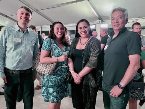 Four people stand smiling together at an indoor event, dressed in semi-formal attire. Other attendees are visible in the background under a marquee ceiling.