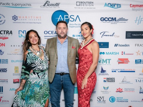 Three people pose and smile in front of a backdrop with various company logos. Two women in floral dresses stand on either side of a man in a suit jacket and blue shirt. All wear leis and look at the camera.