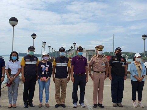 A group of eight people, some in uniforms and some in casual clothes, stand in a row outdoors on a concrete pavement with lamp posts and a partly cloudy sky in the background. All are wearing face masks.