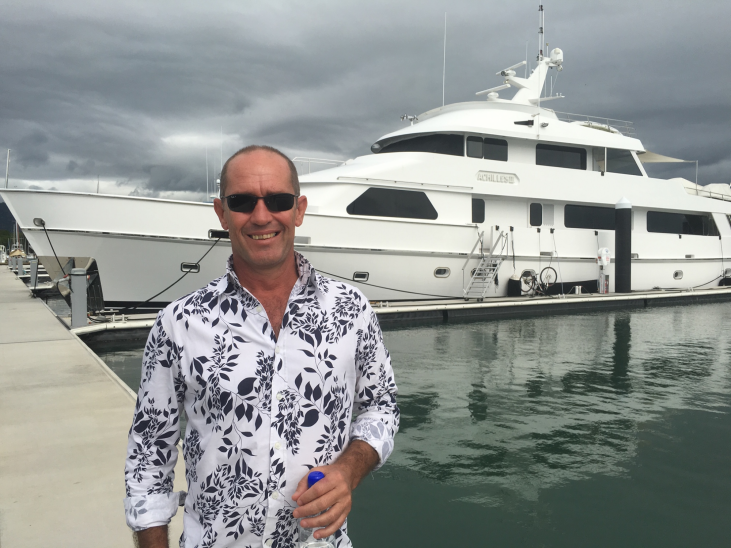 A man in sunglasses and a patterned shirt stands smiling on a quay in front of a large white yacht, with cloudy skies overhead and calm water reflecting the boat.