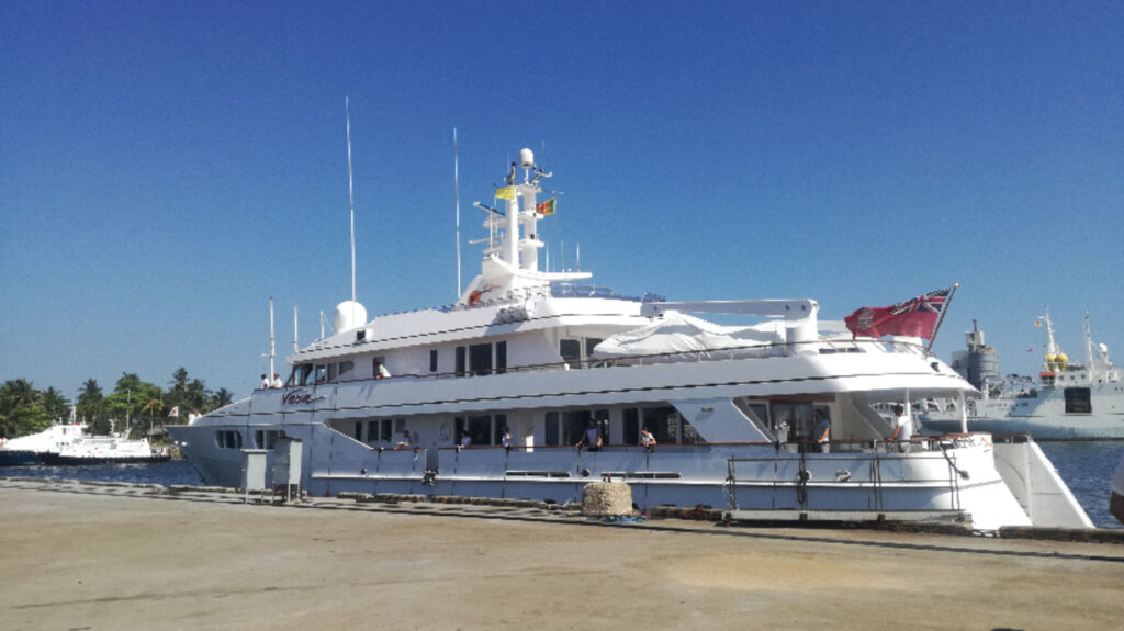 A large white yacht is moored at a harbour under a clear blue sky, with a pier in the foreground and palm trees and other boats visible in the background.
