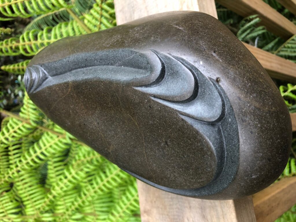 Smooth, dark stone sculpture with elegant, flowing carved lines rests on a wooden surface, surrounded by bright green fern leaves in the background.