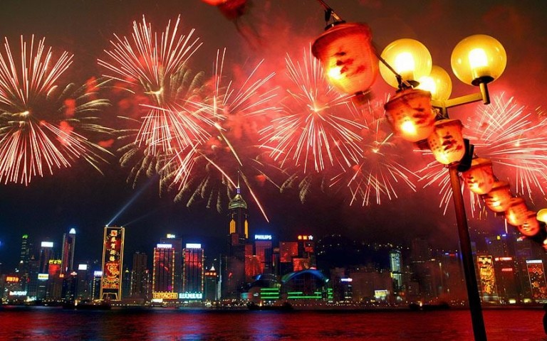 A vibrant city skyline at night is illuminated by colourful fireworks and glowing lanterns, with reflections on the water in the foreground.