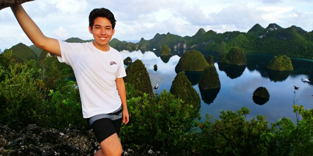 A young man in a white shirt and black shorts smiles whilst standing on a hilltop with lush greenery, overlooking a scenic view of blue water and small, wooded islands.