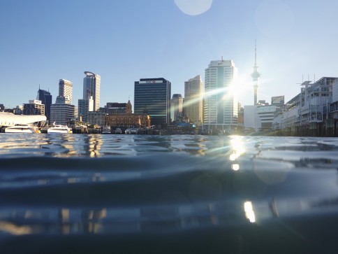 City skyline with modern buildings and a tall tower by the waterfront, sunlight reflecting off the windows and water, with gentle waves in the foreground under a clear blue sky.