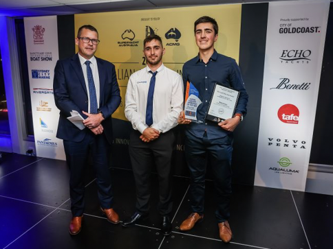 Three men pose on a stage at an awards event. The man on the right holds a trophy and a certificate. A backdrop with sponsor logos and event branding is visible behind them. All three are dressed formally.