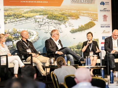 Five panellists sit on stage discussing sustainability at a conference. Four men and one woman are seated with microphones, notepads, and water bottles, with an audience watching and a marina backdrop behind them.