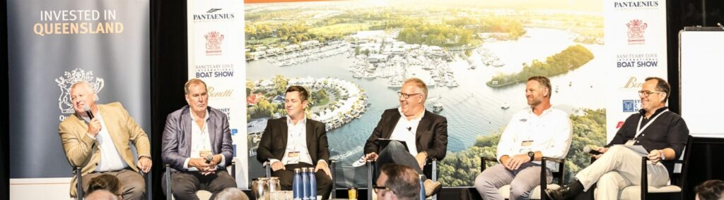 Five men sit on a panel stage speaking at an event, with a backdrop showing a marina and Invested in Queensland signage. The audience is visible in the foreground.