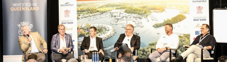 Five men in business attire sit on a panel stage in front of a backdrop showing a marina and trees, with Invested in Queensland signage visible on the left. Bottled water and microphones are on the table in front of them.