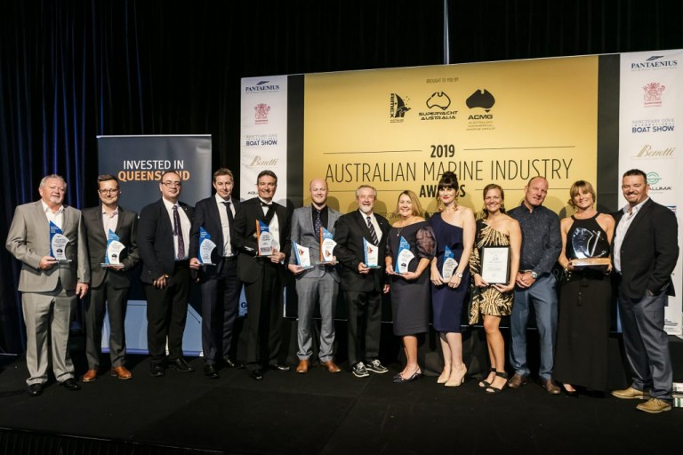 A group of people in formal attire stand on stage holding trophies and plaques at the 2019 Australian Marine Industry Awards, with a large event banner behind them.