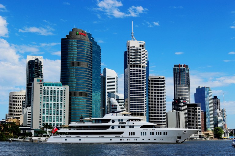 A large white yacht is moored on a river in front of a modern city skyline with tall glass skyscrapers under a bright blue sky with scattered clouds.