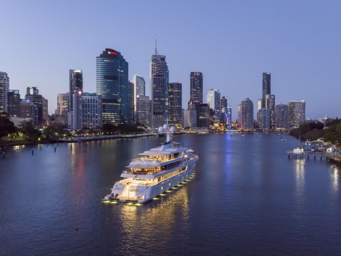 A large yacht with glowing lights sails on a river at dusk, approaching a city skyline filled with tall, modern skyscrapers and illuminated buildings.