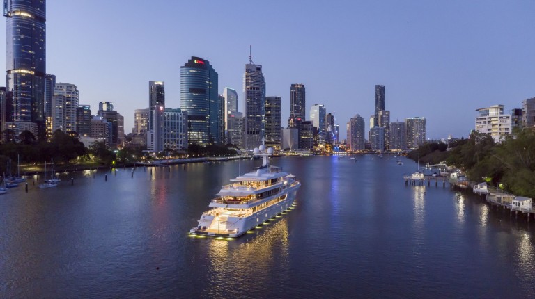 A luxury yacht with lights on sails along a river at dusk, surrounded by high-rise buildings and city lights reflecting in the water, with trees and smaller boats on the riverbank.